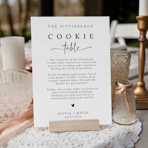 May include: A white card with black text that reads "THE PITTSBURGH COOKIE table." The card is held by a wooden stand. The text describes the tradition of the Pittsburgh cookie table at weddings.