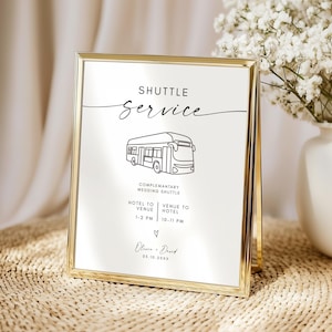 Puede incluir: Un letrero con marco dorado con las palabras "SHUTTLE SERVICE" en escritura negra. Debajo hay un dibujo lineal de un autobús, con el texto "COMPLEMENTARY WEDDING SHUTTLE". El letrero incluye horarios y nombres, y está sobre una superficie tejida.