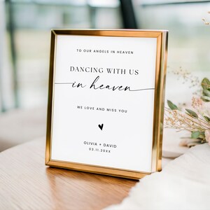 May include: A framed sign with gold trim and white background. The sign reads "TO OUR ANGELS IN HEAVEN", "DANCING WITH US in heaven", and "WE LOVE AND MISS YOU". Below the text is a black heart and the names "OLIVIA + DAVID" with a date.