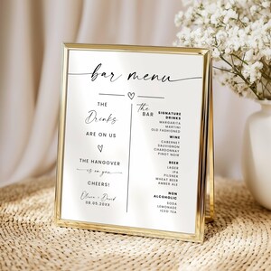 May include: A gold-framed bar menu with black text on a white background. The menu lists signature drinks, wine, beer, and non-alcoholic beverages. The text includes "The Drinks are on us" and "Cheers!" with a wedding date.
