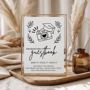 May include: A framed graduation guestbook sign with a gold frame. The sign features a camera illustration with a graduation cap and text that reads "Graduation Guestbook". The sign is on a wooden tray.
