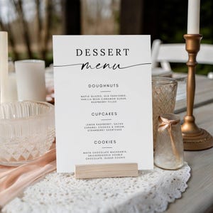 May include: A white dessert menu card with the word "DESSERT menu" in elegant script. The menu lists doughnuts, cupcakes, and cookies with various flavors. The card is held by a wooden stand on a table with other decorative items.