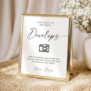 May include: A gold-framed sign with the words "Can't wait to see what Develops" in elegant script. Below is a camera icon and instructions for taking photos. The sign is on a table with flowers in the background.