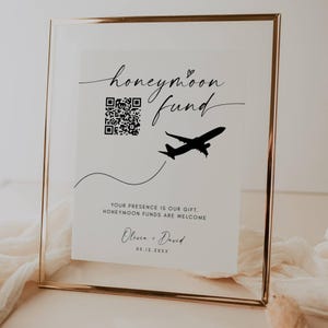 Puede incluir: Un letrero enmarcado con borde dorado y fondo blanco. El letrero dice "honeymoon fund" con un código QR y un gráfico de avión. Debajo del texto está "Your presence is our gift. Honeymoon funds are welcome." Los nombres Olivia y David también están en el letrero.