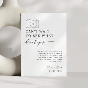May include: A white wedding sign with black text and a camera illustration. The sign reads "Can't wait to see what develops" and includes instructions for guests to take photos. The names "Olivia + David" and a date are also printed.