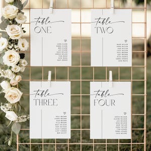 Puede incluir: Cuatro tarjetas de mesa blancas con texto de caligrafía negra, sujetas a una rejilla dorada. Cada tarjeta enumera nombres debajo del número de la mesa. Arreglo floral de rosas blancas y vegetación a la izquierda.