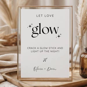 Puede incluir: Un letrero enmarcado en dorado con las palabras "LET LOVE glow" en escritura negra. El letrero también incluye el texto "CRACK A GLOW STICK AND LIGHT UP THE NIGHT!" y los nombres "Olivia + Devin".