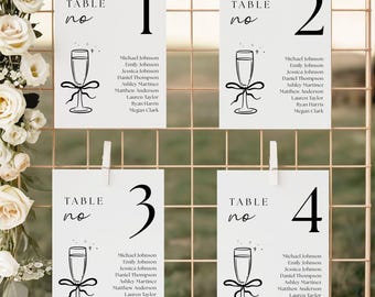 May include: Four white table number cards with black text and a champagne glass illustration. Each card has a number from 1 to 4 and a list of names. The cards are clipped to a gold wire grid.