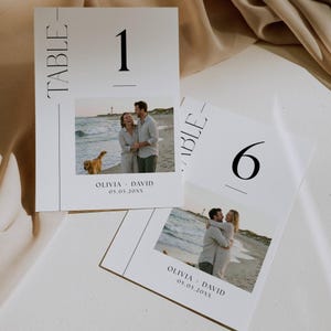 Könnte beinhalten: Zwei weiße Tischkarten mit schwarzem Text und einem Foto. Auf einer Karte steht "TABLE" und "1", auf der anderen "TABLE" und "6". Die Fotos zeigen ein Paar am Strand. Die Karten enthalten auch die Namen "OLIVIA + DAVID" und ein Datum.