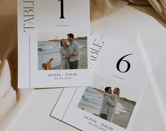 May include: Two white table cards with black text and a photograph. One card reads "TABLE" and "1", the other reads "TABLE" and "6". The photographs show a couple on a beach. The cards also include the names "OLIVIA + DAVID" and a date.