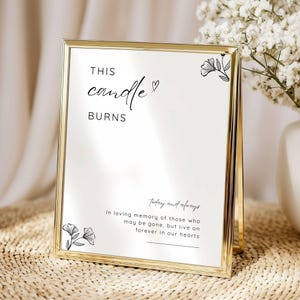 May include: A gold-framed sign with the words "THIS candle BURNS" in elegant script. The sign includes the phrase "today and always" and a memorial message. Floral illustrations adorn the corners. The sign is displayed on a woven surface.