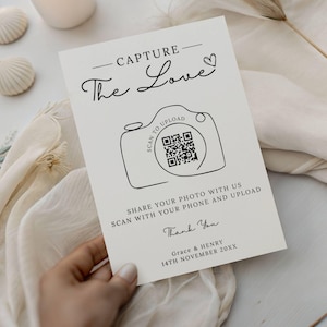 May include: A cream-colored wedding sign with black text that reads "Capture The Love." A camera graphic includes a QR code and the words "Scan to Upload." The sign encourages guests to share photos via phone. Names and a date are at the bottom.