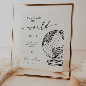 Pode incluir: Uma placa emoldurada com borda dourada apresenta o texto "YOU MEAN THE world TO US" em uma escrita elegante. Abaixo, lê-se "LEAVE YOUR MARK ON OUR WORLD" com uma ilustração de um globo. Os nomes "Olivia + David" e uma data também estão incluídos.