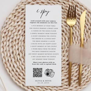 May include: A wedding reception card with the words "i spy" in elegant script. The card lists moments to capture with a camera, along with a QR code and the names Olivia & Devin. A gold fork and knife rest on a beige napkin.