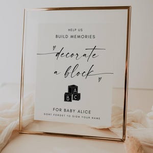 Peut inclure: Un panneau encadré avec une bordure dorée et un fond blanc. Le panneau indique "Help Us Build Memories" et "decorate a block" en écriture élégante. En dessous, un graphique de blocs ABC. Le panneau est personnalisé "For Baby Alice" et invite à signer.