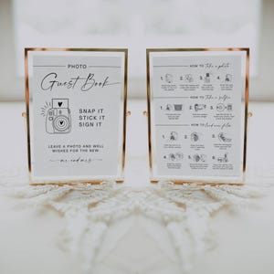 May include: Two framed signs with gold-colored metal frames. The left sign reads "PHOTO Guest Book" with instructions to "SNAP IT STICK IT SIGN IT." The right sign has photo-taking instructions. Both signs are on a white surface.