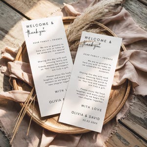 以下が含まれることがあります： 「WELCOME & thank you」とパーソナルメッセージが書かれた、黒い文字の白いウェディングウェルカムカードが2枚。カードは木製のトレイの上に置かれ、ベージュの布とドライフラワーが添えられています。