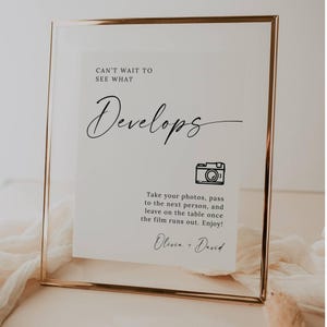 May include: A framed sign with a gold-colored border. The sign has the words "CAN'T WAIT TO SEE WHAT Develops" in black script. Below is a camera icon and instructions for taking photos. The sign is signed "Olivia + David."