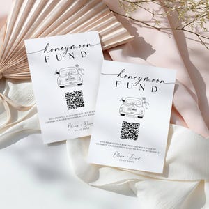 May include: Two white cards with the words "honeymoon fund" in elegant script. Each card features a line drawing of a car with a "Just Married" sign, a QR code, and text. The cards are on a soft, textured surface.