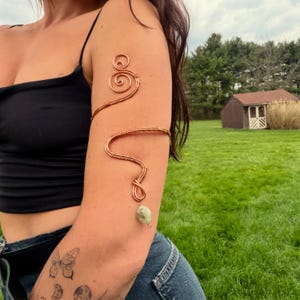 May include: Copper arm cuff with a spiral design and a green stone accent. The cuff wraps around the upper arm and features a flowing, wire-wrapped design. The stone is a light green color and hangs from the lower part of the cuff.