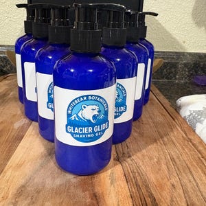 May include: A group of blue bottles of Glacier Glide shaving gel with black pumps. Each bottle has a white label with a bear graphic and the words "Whitebear Botanicals" and "Glacier Glide Shaving Gel". The bottles are arranged on a wooden surface.