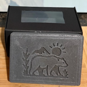 May include: A rectangular, gray soap dish with a carved design featuring a bear, mountains, and a sun. The soap dish is presented in front of a black box with a clear window.