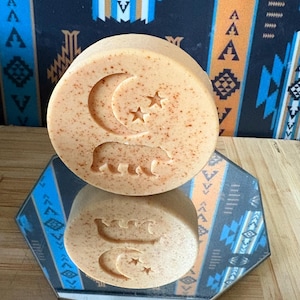 May include: A round, light brown soap bar with a bear, crescent moon, and star design. The soap is reflected in a mirrored surface. The background features a patterned fabric with blue, orange, and white geometric shapes.