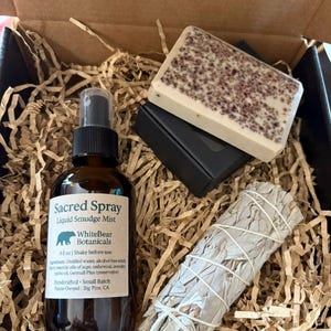 Op de afbeelding: Een geschenkdoos met een bruine glazen fles "Sacred Spray" liquid smudge mist, een stuk zeep en een bundel witte salie. De fles heeft een zwarte sproeikop en een wit etiket. De zeep is rechthoekig met donkere spikkels.