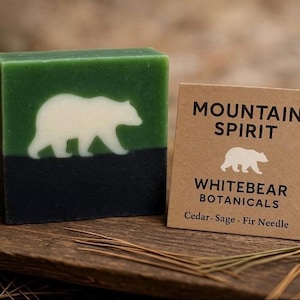 May include: A handcrafted soap bar with a white bear silhouette on a green top and black bottom. A small card reads "MOUNTAIN SPIRIT" and "WHITEBEAR BOTANICALS" with the scent description "Cedar-Sage - Fir Needle".