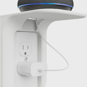 Op de afbeelding: Een witte wandplank met een ingebouwd stopcontact en een slimme luidspreker. De luidspreker is zwart met een blauw licht. Een witte oplader is aangesloten op het stopcontact, met een witte kabel naar de luidspreker.