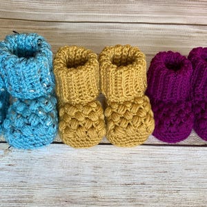 May include: A collection of six handmade baby booties in three colors: light blue, mustard yellow, and deep purple. The booties are crocheted with a textured stitch, showcasing a cozy and handcrafted aesthetic. The booties are arranged in pairs.