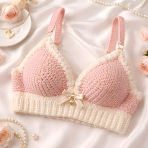 Peut inclure: Soutien-gorge en crochet rose et crème fait à la main, avec des bretelles réglables. Le soutien-gorge présente une bordure festonnée et un petit nœud avec un charme au centre. La maille douce et texturée est présentée sur une surface en satin blanc, avec des accents floraux et perles.