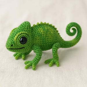 Op de afbeelding: Een handgemaakte gehaakte kameleon in groentinten. Het reptiel heeft een getextureerd lichaam, een gekrulde staart en grote zwarte ogen. De kameleon staat op een witte ondergrond, wat het gedetailleerde vakmanschap benadrukt.