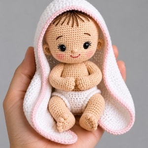 May include: A crocheted baby doll with a white and pink-trimmed hood and diaper. The doll has brown hair, rosy cheeks, and is held in a hand. The doll is made of beige yarn.