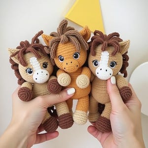 May include: Three crochet horse dolls in shades of tan, brown, and orange. Each doll has a white blaze, dark brown mane and tail, and large, expressive eyes. The dolls are held up by a person's hands.