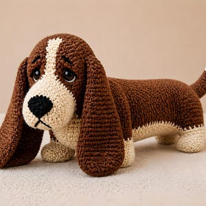 Op de afbeelding: Een gehaakte basset hound hondenspeelgoed in bruin en crème. De hond heeft lange, slappe oren en een trieste uitdrukking. Het lichaam is bruin, met crèmekleurige poten en een witgepunte staart.