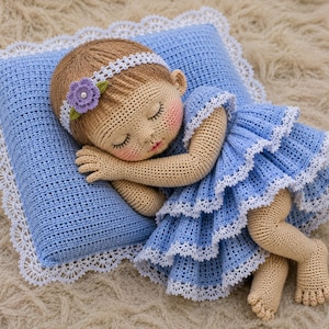 Peut inclure: Une poupée au crochet aux cheveux châtain clair, portant un bandeau blanc avec une fleur violette et une robe bleue à volants avec une bordure en dentelle blanche. La poupée dort sur un oreiller assorti au crochet avec des bords en dentelle.