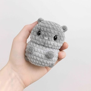 Puede incluir: Un hámster de ganchillo gris hecho a mano con ojos negros, orejas pequeñas y una nariz diminuta. El peluche se sostiene en una mano sobre un fondo blanco. El hámster tiene un aspecto texturizado y suave.