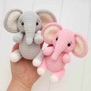 Puede incluir: Dos juguetes de elefante hechos a mano con ganchillo. Uno es gris claro con detalles blancos y crema, el otro es rosa con detalles blancos y crema. Ambos tienen orejas grandes, trompas y ojos negros pequeños. Los juguetes son suaves y tiernos.