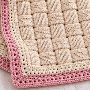 May include: A cream-colored knitted blanket featuring a basketweave pattern. The blanket is finished with a decorative border consisting of a row of cream-colored stitches followed by a wider, textured edge in a soft, dusty pink yarn.