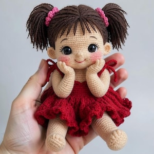 Cute sitting toddler doll crochet pattern PDF featuring an amigurumi girl with brown pigtails, rosy cheeks, large eyes, red ruffled dress with bow straps, hands-on-cheeks pose, detailed crochet doll tutorial and digital download for crocheters.