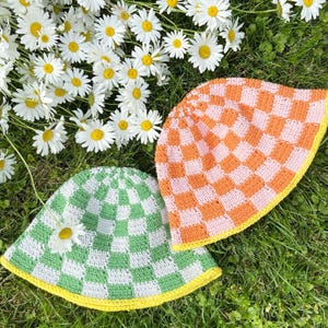 Puede incluir: Dos sombreros de pescador de ganchillo con un patrón de tablero de ajedrez. Un sombrero es verde y blanco, el otro es naranja y rosa. Ambos sombreros tienen un borde amarillo. Las margaritas están esparcidas alrededor de los sombreros sobre hierba verde.