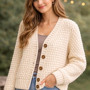 Puede incluir: Cárdigan de punto texturizado color crema con cuello en V y cierre de botones. El cárdigan tiene dobladillo y puños acanalados, y se combina con una camiseta neutra y vaqueros azules. Los botones son de color marrón cálido.