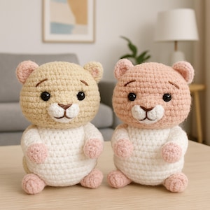May include: Two handmade crochet bear toys. One bear is tan and white, the other is pink and white. Both have black eyes, brown noses, and pink paws. The bears are sitting on a light brown surface.