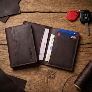 May include: A brown leather wallet, open to display cards and cash. The wallet is on a wooden surface, with a separate cardholder and a car key fob nearby. The wallet has the brand name "ROBINS" embossed on the front.