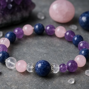 Könnte beinhalten: Ein Armband mit Edelsteinperlen. Das Armband enthält rosa, lila, blaue und klare Perlen. Zusätzliche Edelsteine und Kristalle sind auf einer grauen Oberfläche verstreut. Das Armband ist ein Schmuckstück.