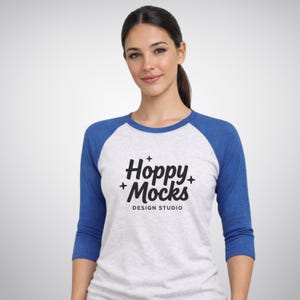 Op de afbeelding: Een baseball-stijl shirt met een witte body en blauwe mouwen. Het shirt toont de tekst "Hoppy + Mocks DESIGN STUDIO" in het zwart. Het model draagt het shirt.