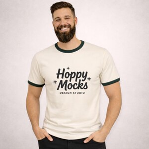 Op de afbeelding: Crèmekleurig ringer t-shirt met donkergroene randen rond de kraag en mouwen. Het shirt heeft de tekst "Hoppy Mocks DESIGN STUDIO" in zwart. De persoon draagt een zwarte broek.