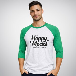 Op de afbeelding: Een wit en gemêleerd honkbal-T-shirt met groene mouwen. Het shirt heeft de tekst "Hoppy + Mocks DESIGN STUDIO" in zwart. Het shirt heeft driekwart mouwen.