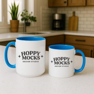 May include: Two white ceramic mugs with blue interiors and handles. The mugs have the text "HOPPY MOCKS DESIGN STUDIO" printed in black. One mug is larger than the other.
